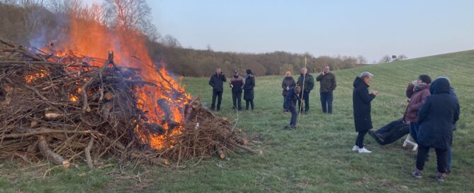 Ein großes Feuer brennt auf einem Feld und menschen stehen außen rum und unterhalten sich und trinken heiße Getränke.
