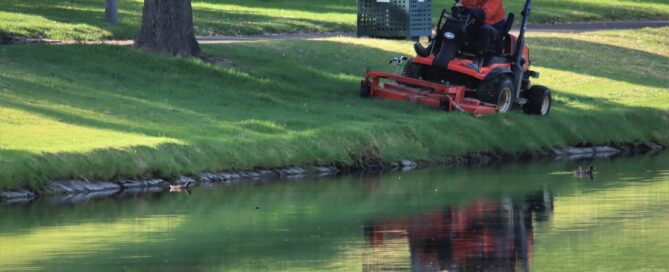Ein großer Aufsitzrasenmäher mäht an der Kante eines Gewässers den Rasen.
